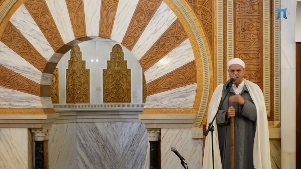 Jutba junto al gran arco de herradura del mimbar, con el imam de chilaba gris ante el micrófono