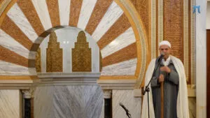 Jutba junto al gran arco de herradura del mimbar, con el imam de chilaba gris ante el micrófono