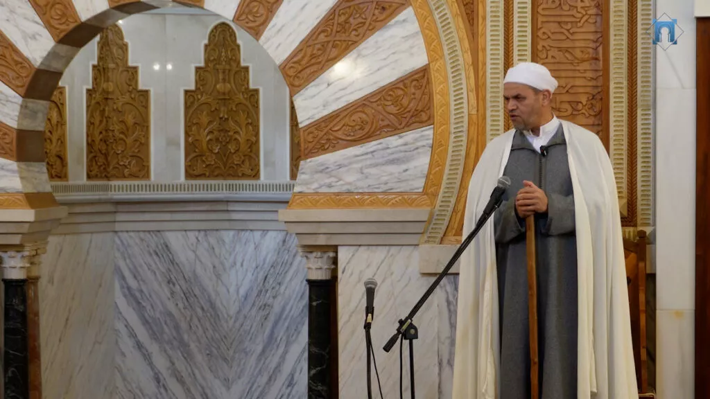 Imam de chilaba gris y capa blanca, apoyado en el bastón, hablando ante los micrófonos del mimbar