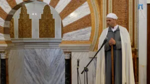 Imam de chilaba gris y capa blanca, apoyado en el bastón, hablando ante los micrófonos del mimbar