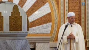 Jatib con turbante blanco y chilaba color crema da la jutba junto al arco labrado del mihrab