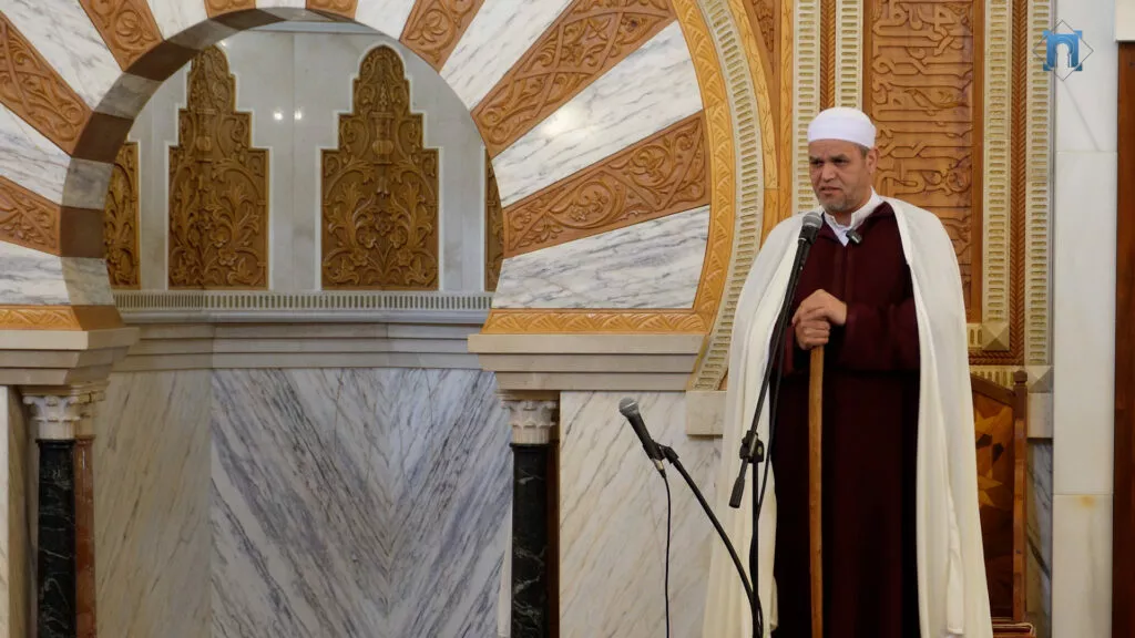 Imam de chilaba granate y capa blanca dando la jutba desde el mimbar de mármol tallado