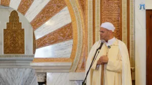 Imam con chilaba y capa color crema durante la jutba, junto al arco tallado del mimbar