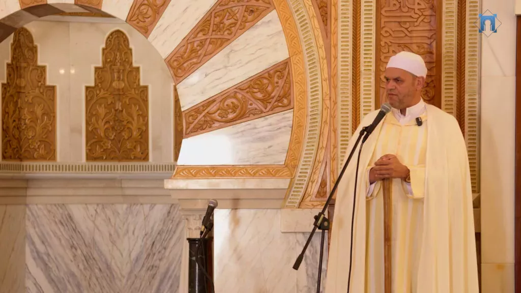 Imam con manto claro y bastón da el sermón junto al arco tallado del mihrab