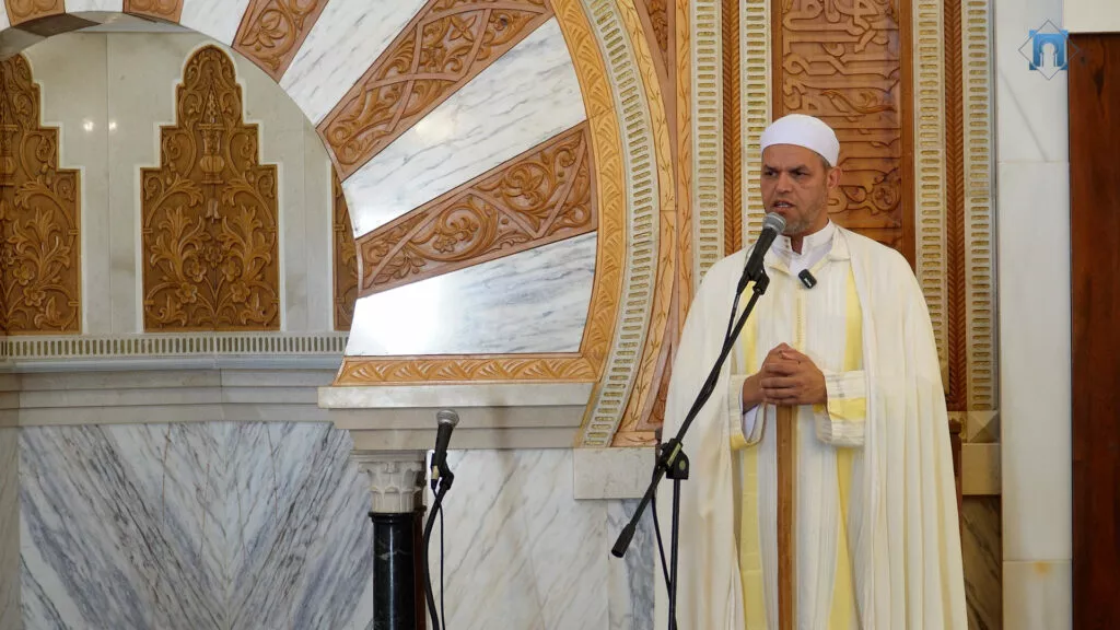 Imam de chilaba clara con franja dorada dando la jutba ante los micrófonos del mimbar