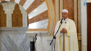 Imam de chilaba clara con franja dorada dando la jutba ante los micrófonos del mimbar
