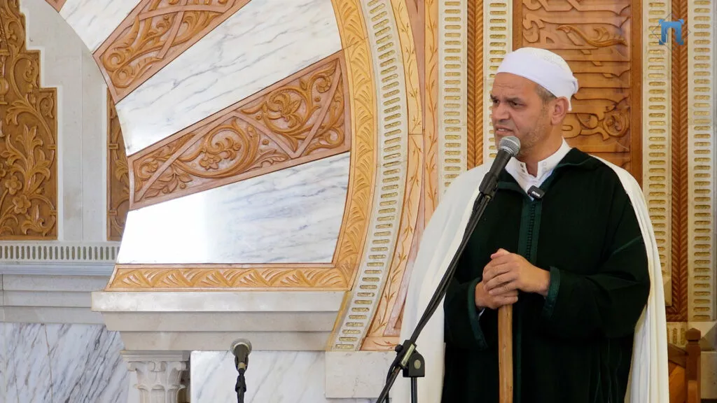 Imam de chilaba verde oscura y capa blanca dando la jutba apoyado en el bastón