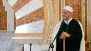 Imam de chilaba verde oscura y capa blanca dando la jutba apoyado en el bastón